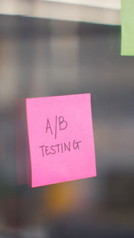 Vertical-Video-Close-Up-Of-Woman-Putting-Sticky-Note-With-A/B-Testing-Written-On-It-Onto-Transparent-Screen-In-Office